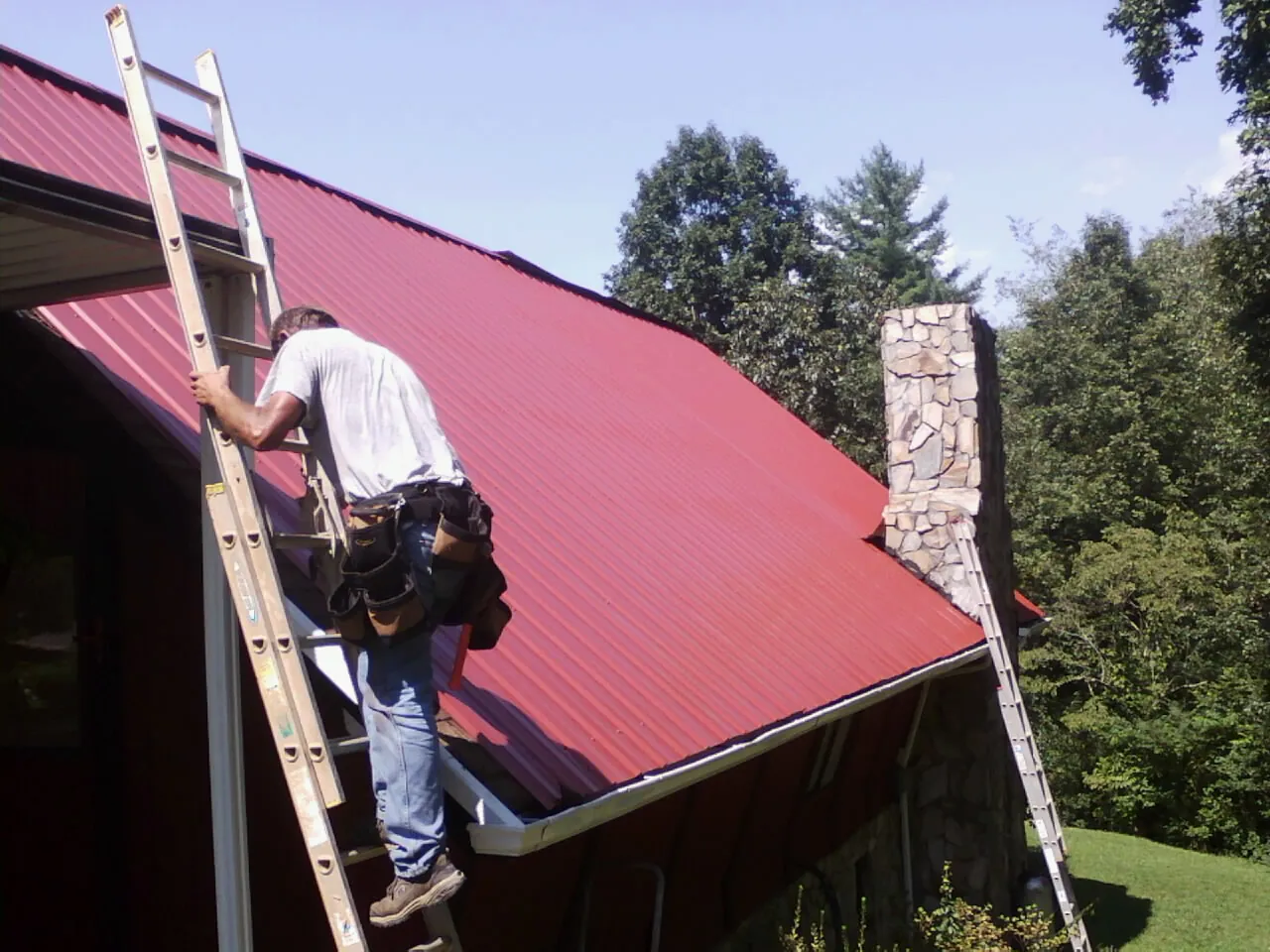 Roofer on a ladder beside a red metal roof and stone chimney — Soffit & Fascia Repair by Roofing Brothers in Nyack